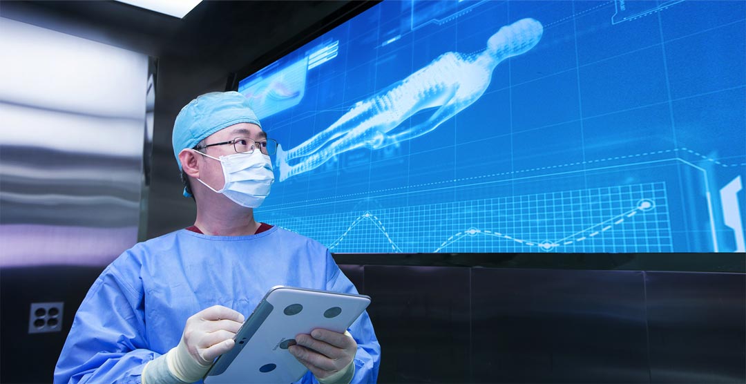 A surgeon in an operating room looking at a large digital screen displaying a 3D model of a human body, holding a tablet.
