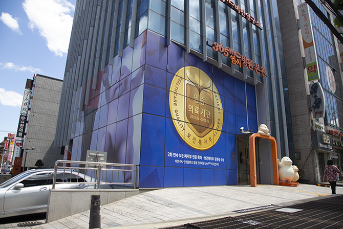 The entrance of a modern building with a large gold seal displayed on a blue panel, alongside glass windows and company branding above the doorway.